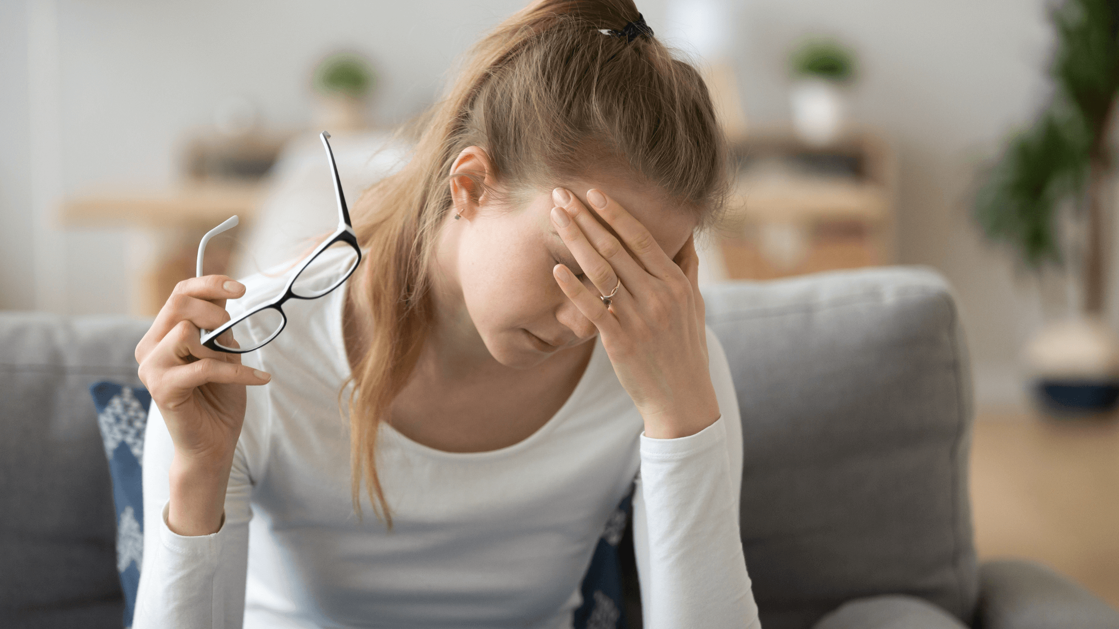 Woman holding her head and a pair of glasses, with a blurry background, symbolizing blurry vision and eye strain from cataract warning signs.