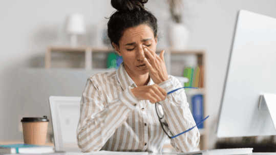 A woman sitting at a desk rubbing her irritated eyes and holding her glasses, illustrating the discomfort caused by chronic dry eye syndrome in the winter.