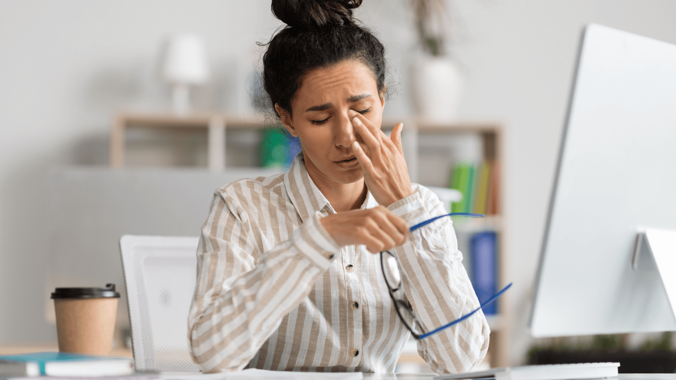 A woman sitting at a desk rubbing her irritated eyes and holding her glasses, illustrating the discomfort caused by chronic dry eye syndrome in the winter.