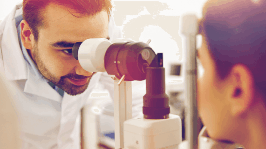 An optometrist looking into a patient's eye with a slit lamp, symbolizing the detailed examination process that can now be enhanced or replaced by comfortable Optomap retinal imaging.