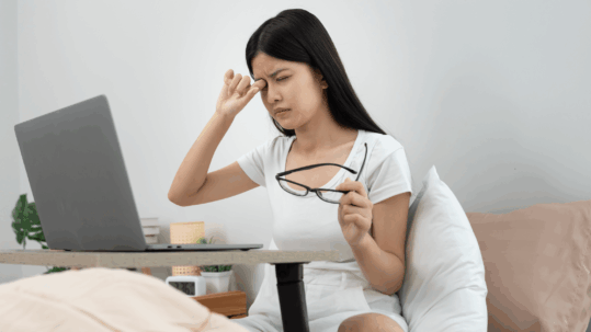 A young woman, likely working on a laptop, holds her hand over her eye in discomfort while holding her glasses, symbolizing eye strain or the distraction caused by new floaters and flashes.