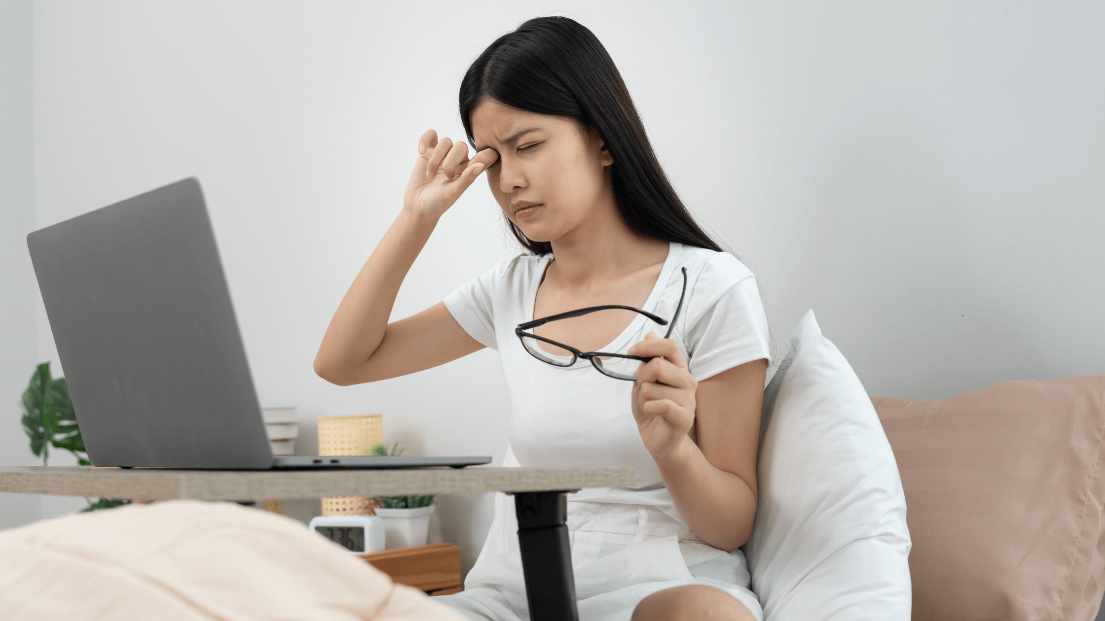 A young woman, likely working on a laptop, holds her hand over her eye in discomfort while holding her glasses, symbolizing eye strain or the distraction caused by new floaters and flashes.