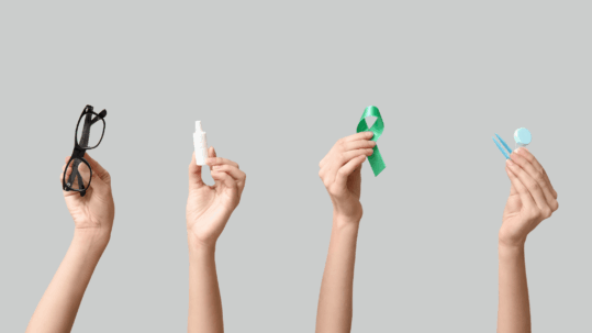 Four hands holding objects related to eye care and Glaucoma awareness: a pair of black-rimmed glasses, a bottle of prescription eye drops, a green Glaucoma awareness ribbon, and instruments for an eye exam.