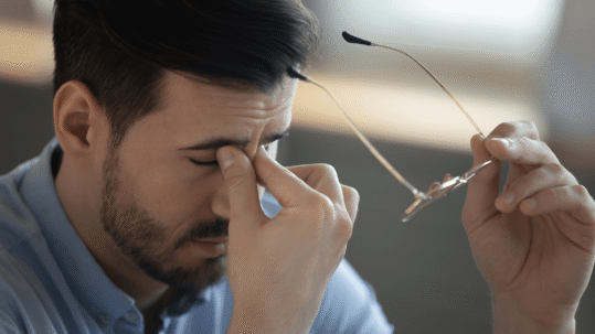 A man holding his glasses and rubbing the bridge of his nose due to eye strain or blurry vision.